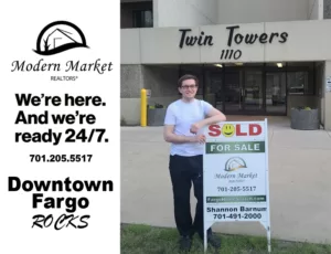 A Modern Market REALTORS® agent stands in front of the Twin Towers building in downtown Fargo, holding a 'SOLD' sign displaying the Modern Market REALTORS® logo and agent contact information