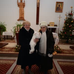 Two elderly women stand in a decorated Polish church corner, holding a baby in white winter attire. A religious statue and icons are visible in the background, capturing the heartwarming essence of Christmas.