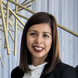 Smiling woman with straight shoulder-length hair wearing a black blazer and white blouse, standing in front of a modern decorative backdrop.