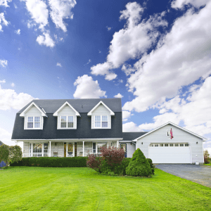 Beautiful two-story home with a well-manicured green lawn, attached garage, and a spacious front porch under a bright blue sky. A residential home for sale in a suburban neighborhood