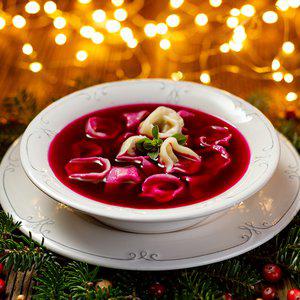 A white bowl of red soup containing dumplings, garnished with a sprig of green herb, is placed on a white plate in the cozy corner of a room, softly illuminated by festive Christmas lights.