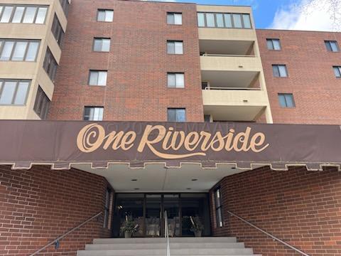 Front entrance of the One Riverside condominium building in Moorhead, MN, featuring a brick facade, signature awning with building name, and a set of stairs leading up to the lobby doors.