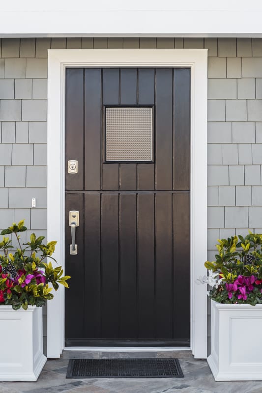 Modern black front door with a small window, white trim, gray shingle siding, and potted flowers on each side.