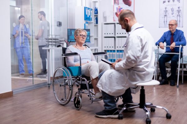 A medical professional sits on a stool, consulting with a person sat in a wheelchair.