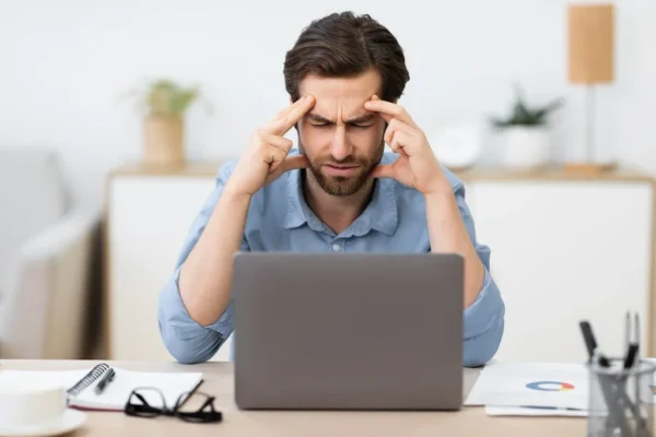 A man is sitting, facing a laptop. He has his hands pressing against his head. He appears to be suffering from head pain.