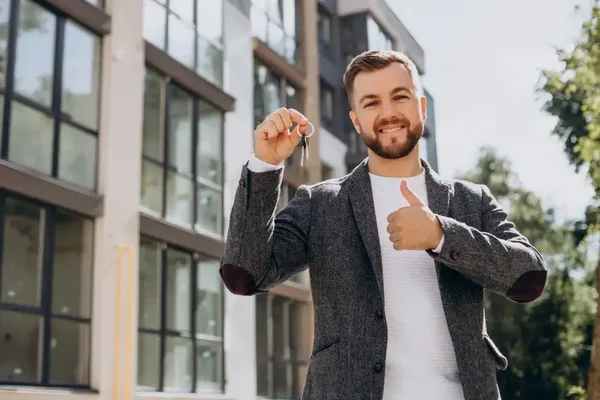 Young man celebrates achievement with keys, smiling confidently outside a modern building.