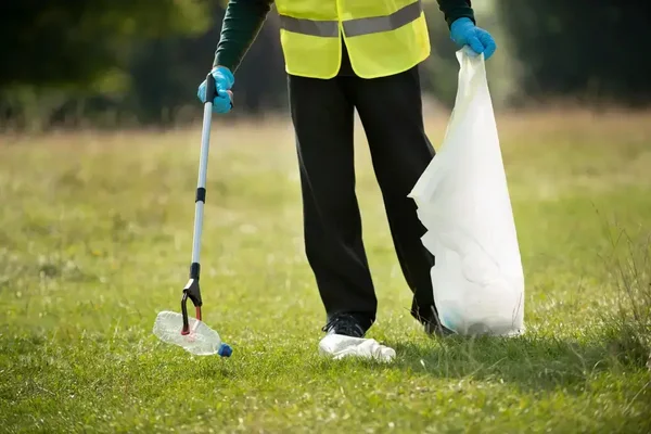 Person in a yellow vest cleans litter in a sunny park, promoting environmental stewardship.