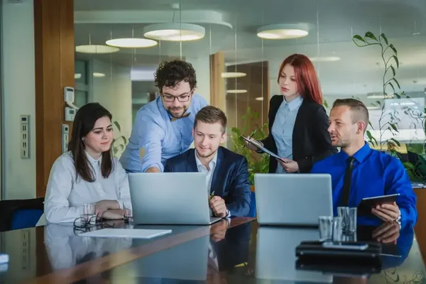 Professionals collaborate around a laptop in a bright, modern office meeting.
