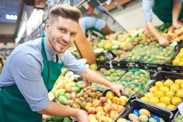 Cheerful employee interacts with colorful fresh fruits, creating a welcoming grocery shopping experience.