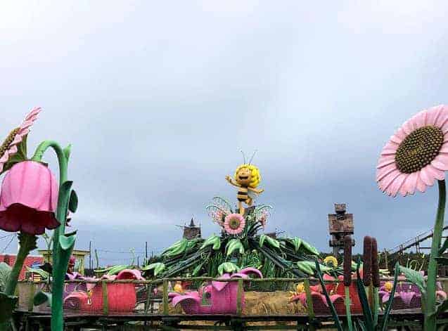 Colorful outdoor amusement park ride with large flower decorations and a smiling bee character on top awaits at Majaland Amusement Park Kownaty, under a cloudy sky.