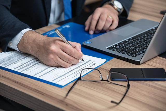A person writing on a form while working on a laptop computer