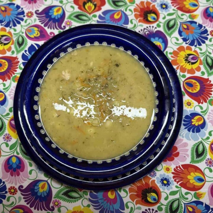 A bowl of creamy Ogorkowa soup with herbs and small vegetable pieces, placed on a blue plate. The background is a colorful, floral-patterned tablecloth, evoking the charm of a traditional Polish recipe.