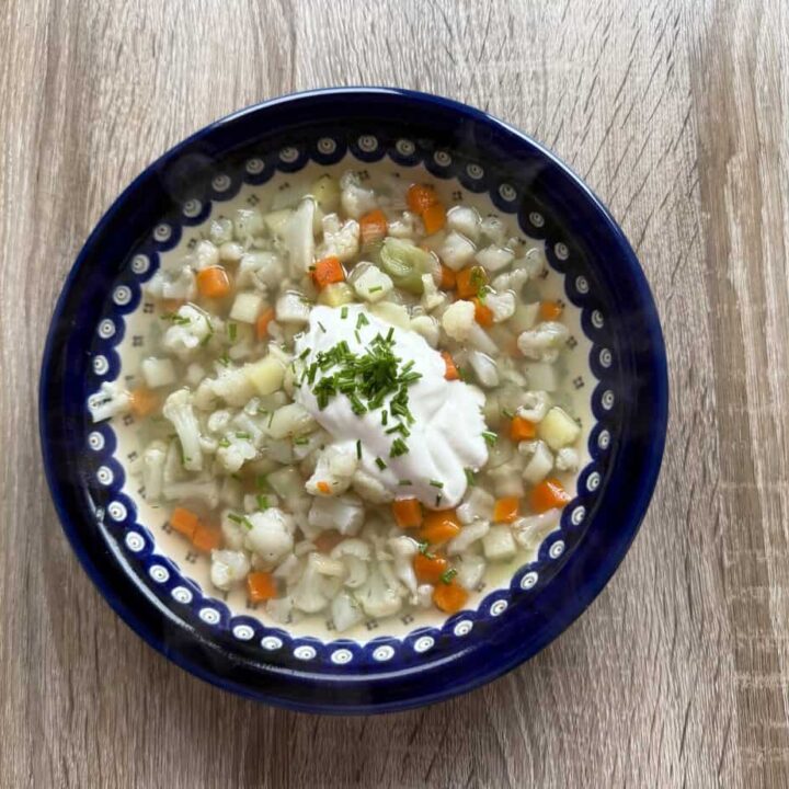 A bowl of creamy Polish Cauliflower Soup with diced carrots, potatoes, and herbs, topped with a dollop of sour cream and chopped chives, on a wooden table.