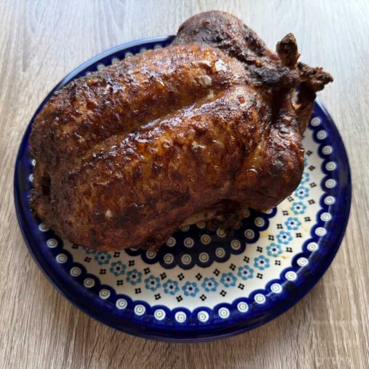A whole roasted chicken with crispy, browned skin sits on a patterned blue and white plate on a wooden surface, reminiscent of a classic Polish duck recipe presentation.