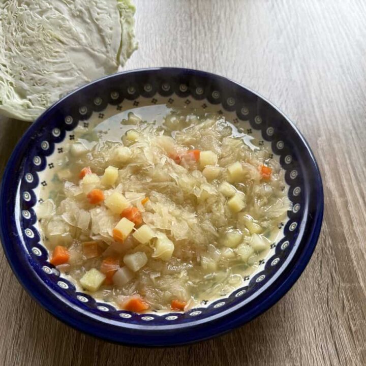 A bowl of Vegetarian Kapusniak, filled with diced potatoes and carrots, sits on a wooden table next to a half head of cabbage.