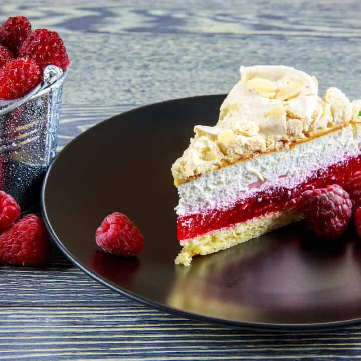 A piece of cake with raspberries on a black plate, featuring a malinowa chmurka recipe.