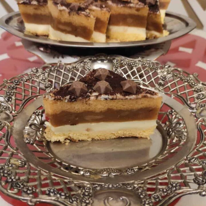 A slice of caramel and chocolate layered cake on an ornate silver plate with more slices of the layered apple cake in the background.