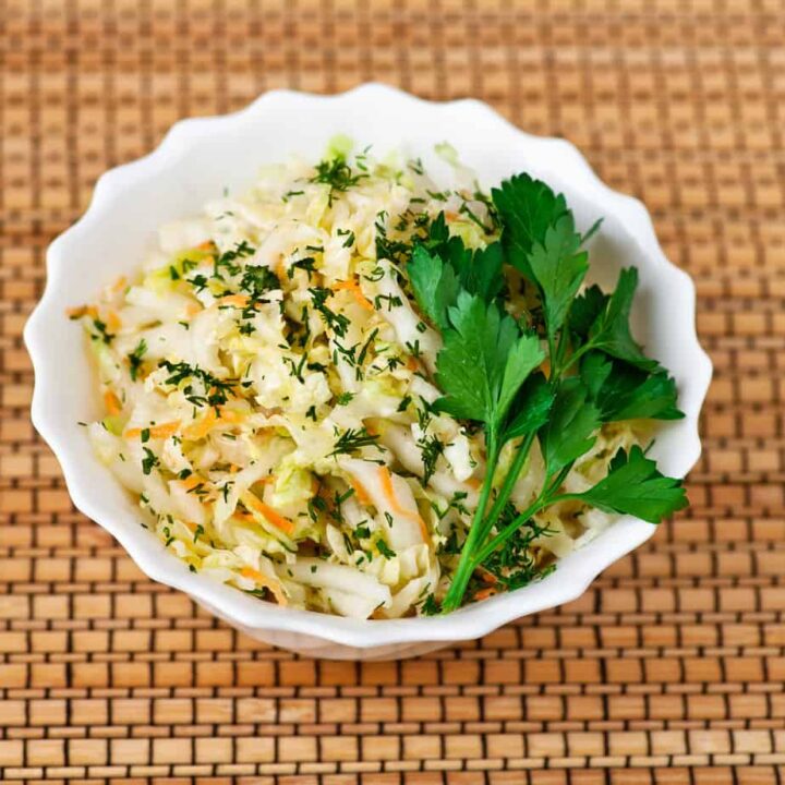 A white bowl filled with Polish sauerkraut salad, garnished with fresh parsley, placed on a woven mat.