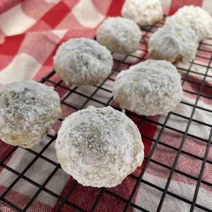 Several powdered sugar-coated Polish snowball cookies are cooling on a black wire rack over a red and white checkered cloth.