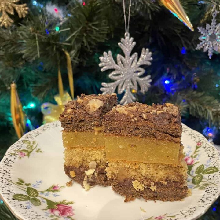 A floral plate holds two square pieces of Walnut Layered Cake in front of a decorated Christmas tree with gold and silver ornaments.