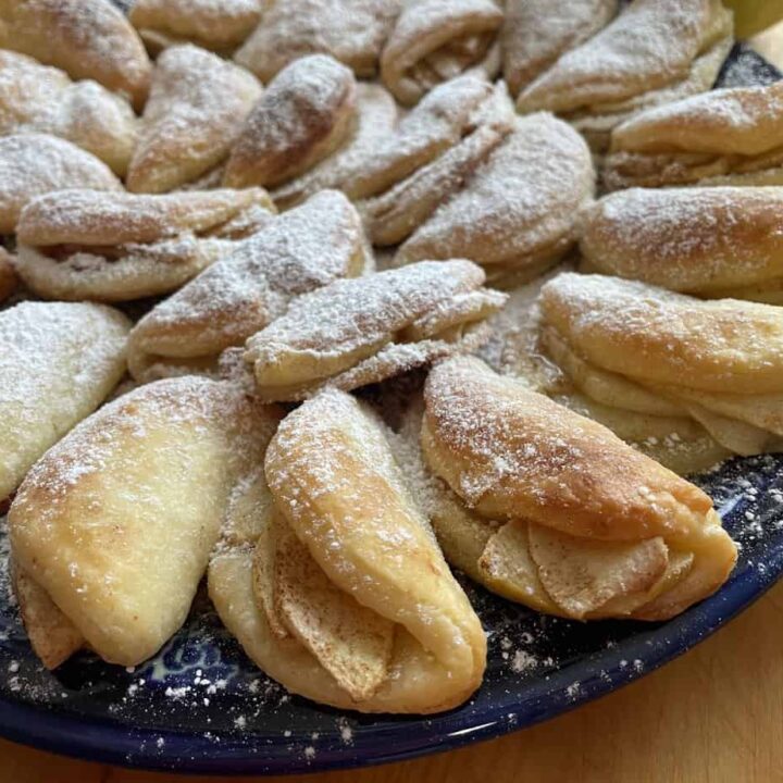 A plate of baked, crescent-shaped Polish Apple Cookies dusted with powdered sugar is displayed on a wooden surface.