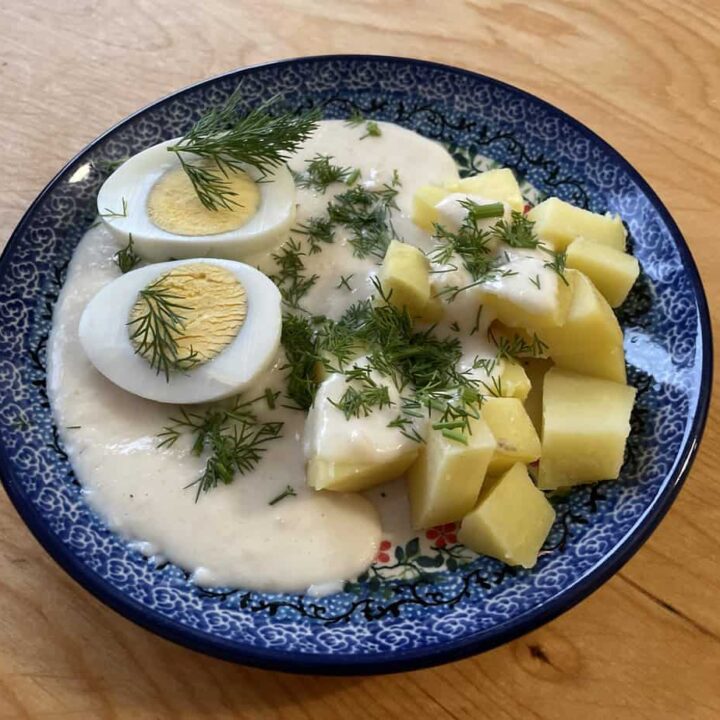 A blue-patterned bowl containing Polish Eggs In Horseradish Sauce with halved boiled eggs, cut potatoes, creamy white sauce, and dill garnish, placed on a wooden surface.