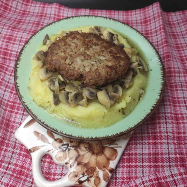 A cooked patty sits atop mashed potatoes and sliced mushrooms on a green plate with a red and white checkered cloth underneath, reminiscent of a classic Polish Bryzol Recipe from the PRL Era.