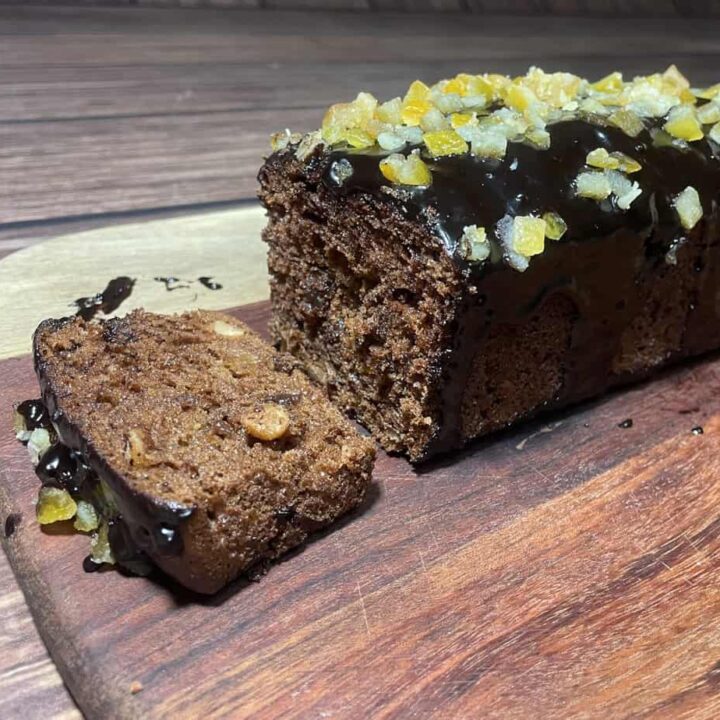 Loaf of chocolate gingerbread cake with nuts and candied fruit on top, partially sliced, on a wooden cutting board.