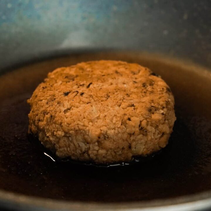 A brown lentil patty is sizzling in a pan with hot oil, creating a delicious aroma.