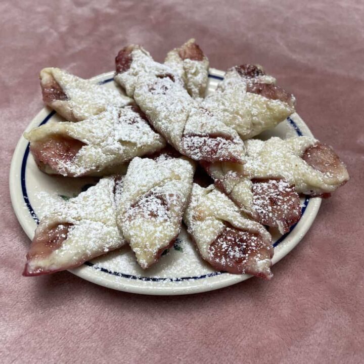 A plate of star-shaped Smalc&oacute;wki, Polish Lard Cookies dusted with powdered sugar, filled with pink jam, resting on a soft pink surface.