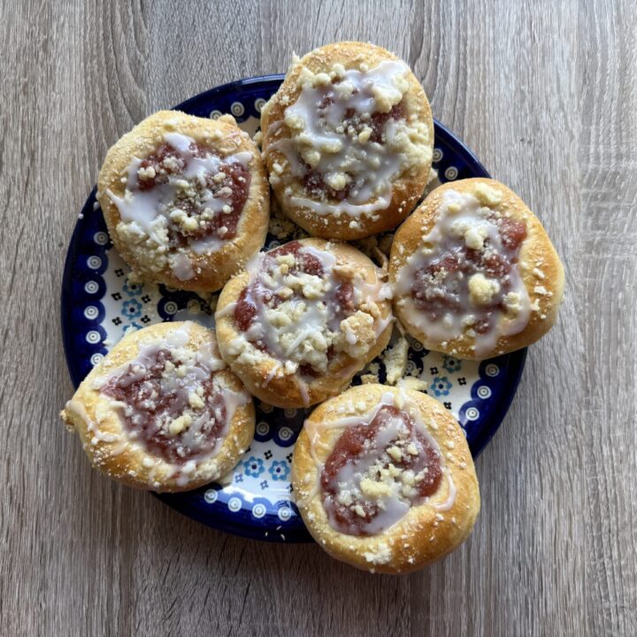 A plate of six round Drożdż&oacute;wki&mdash;Polish Apple Yeast Buns&mdash;with fruit filling, glaze, and crumb topping, arranged on a patterned blue dish on a wooden surface. An easy recipe for a classic treat.