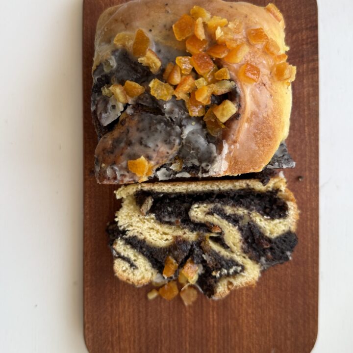 A glazed sweet bread roll, reminiscent of classic Makowiec, with a dark poppyseed cake filling and chopped orange candied fruit on top, sliced and served on a small wooden board.