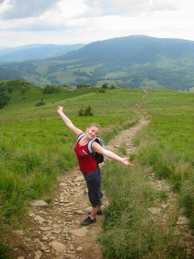 A person with a backpack stands on a rocky trail in a grassy, hilly landscape, arms outstretched, with mountains and clouds in the background—perfect inspiration for anyone considering solo travel Poland.