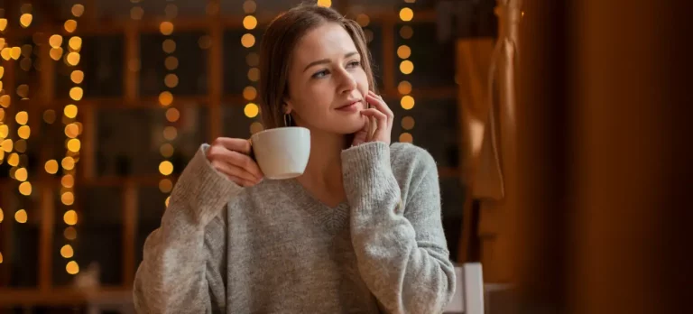 Eine Frau sitzt entspannt mit einer Tasse Kaffee und blickt verträumt, ein Moment der Selbstreflexion beim allein sein lernen.