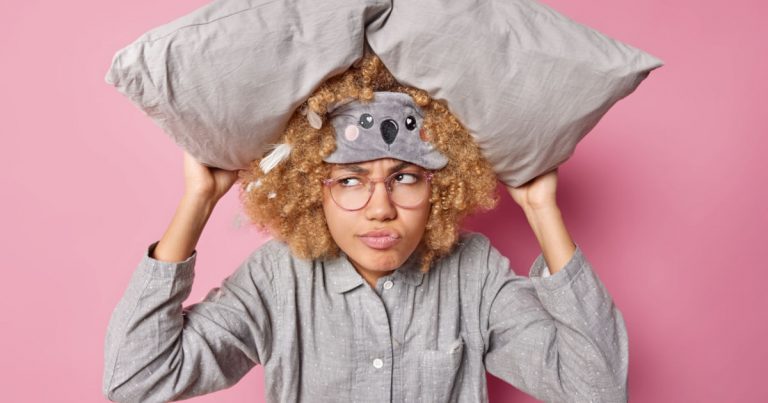 Frau hält sich ein Kissen über den Kopf, trägt eine Schlafmaske und wirkt genervt – typische Zeichen von Schlafproblemen durch saisonale Depression.