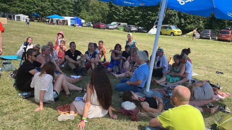 A group of people sit in a circle on the grass under a large umbrella at an outdoor event, with tents and parked cars in the background.