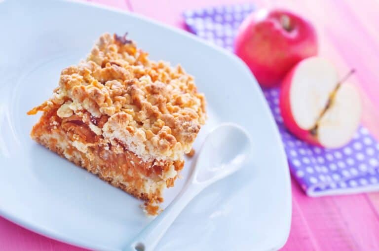 A slice of Polish apple cake with meringue on a plate with a spoon, accompanied by a whole apple and a half-cut apple in the background.