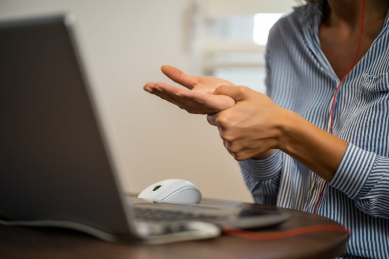 The hands of someone sat near a laptop computer. They are pushing their thumb from one hand in to the palm of their other hand.