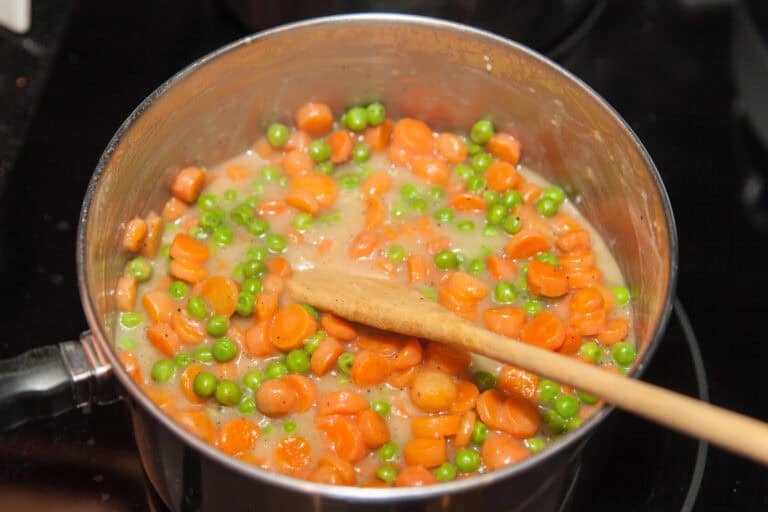 A wooden spoon stirs a pot of Polish creamed peas and carrots salad, with each swirl blending the cooked peas and sliced carrots in a smooth, creamy sauce.