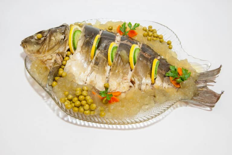 Polish carp in aspic is elegantly presented with lemon slices, lettuce, peas, and diced vegetables on a clear glass plate.