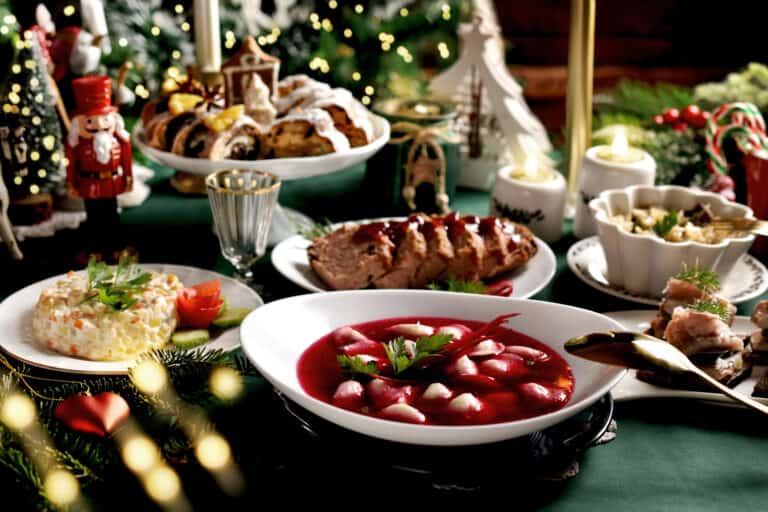 A table set for a festive meal with 12 Polish Christmas dishes including red soup, roasted meat, salad, and pastries, adorned with holiday decorations such as candles, a nutcracker figurine, and a Christmas tree in the background.