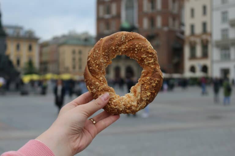 A person holding a Polish bagel with a bite taken out of it in front of a blurry urban background.