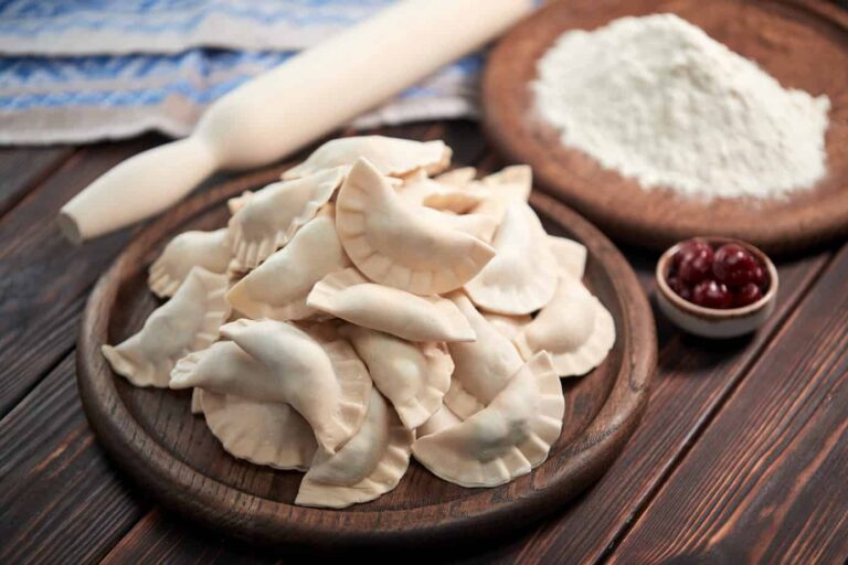 A wooden platter filled with uncooked pierogi, next to a small bowl of cherries and a plate of flour, evokes the question: where did pierogi originate from? A rolling pin and a blue-and-white cloth lie in the background.