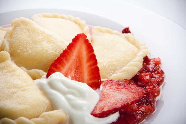 A close-up of a plate featuring Polish pierogi served with fresh sliced strawberries, a dollop of white cream, and a drizzle of strawberry puree.
