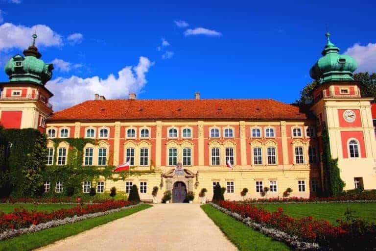 A large Baroque-style palace with two green domed towers, a red roof, and manicured gardens under a blue sky&mdash;just one of the many reasons to visit Poland.