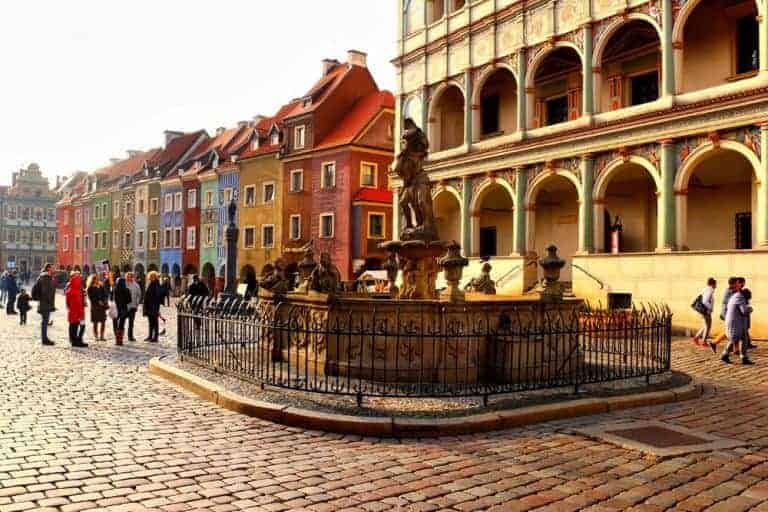 A stone fountain surrounded by an iron fence sits in a cobblestone square with colorful buildings and people walking nearby—an example of the interesting Poznan sights found throughout the city.