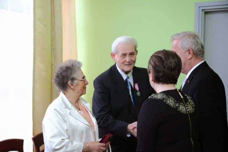 Four older adults, two men and two women, stand indoors talking and shaking hands; one man wears a medal on his suit, perhaps sharing Poland facts or swapping funny Poland trivia as they enjoy each other's company.