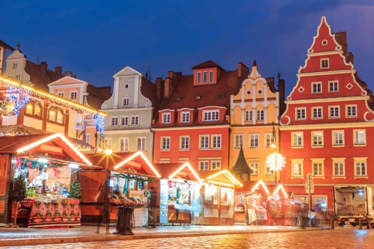 Colorful historic buildings line a street behind illuminated Christmas market stalls at dusk, with lights and decorations visible in the evening scene.