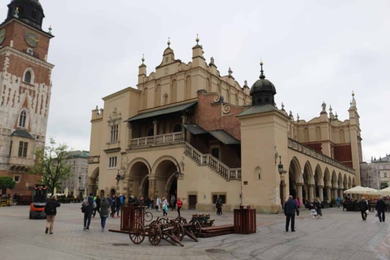 People walk around the exterior of a historic building with arches and towers in a city square; old cannons are displayed in the foreground.