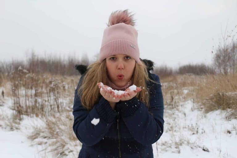 A person in a pink hat and blue coat stands outside in a snowy field, holding snow in their hands and blowing on it.
